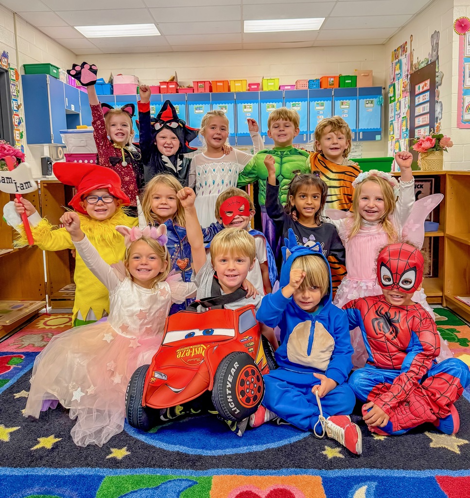 A group of pre-kindergarten students pose cheerfully in a colorful classroom, wearing a variety of costumes including superheroes, princesses, animals, and storybook characters. They raise their arms excitedly while smiling for the camera.