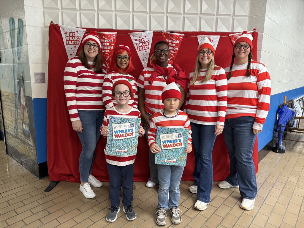 Six teachers and two students dressed as “Where’s Waldo” characters pose in front of a red backdrop that reads “Be Drug Free.” Everyone is wearing red-and-white striped shirts, round glasses, and red hats. Two children hold Where’s Waldo books.