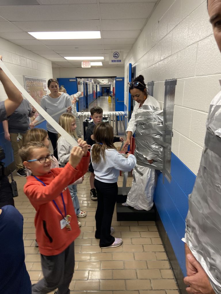 Children and teachers in a school hallway help tape an adult to the wall with strips of duct tape during a fun school activity.