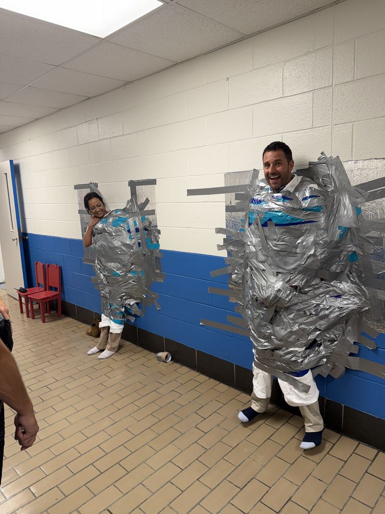 Two adults are fully taped to a blue-and-white hallway wall, smiling and laughing as students watch the result of the duct tape challenge.