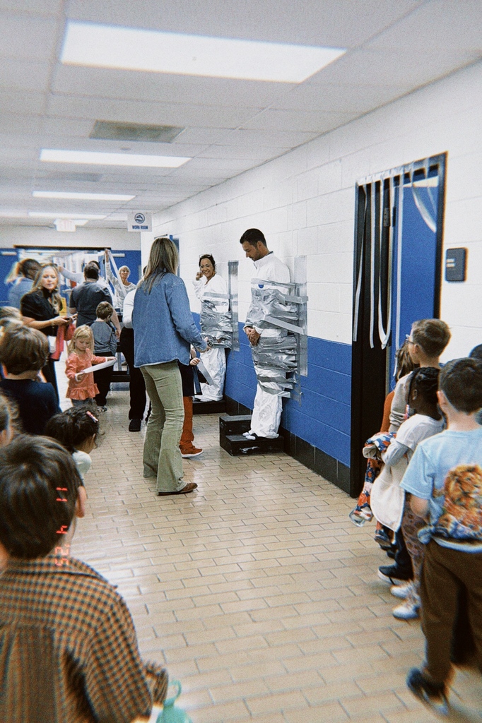 A group of children and teachers gather in the hallway watching two adults taped to the wall, smiling and laughing during the event.