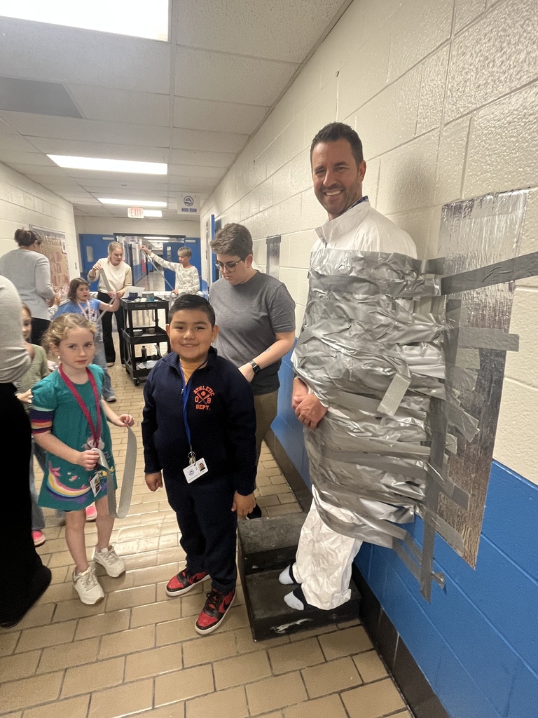 A smiling adult stands taped to the wall as students pose nearby, showing excitement during the duct tape challenge event.