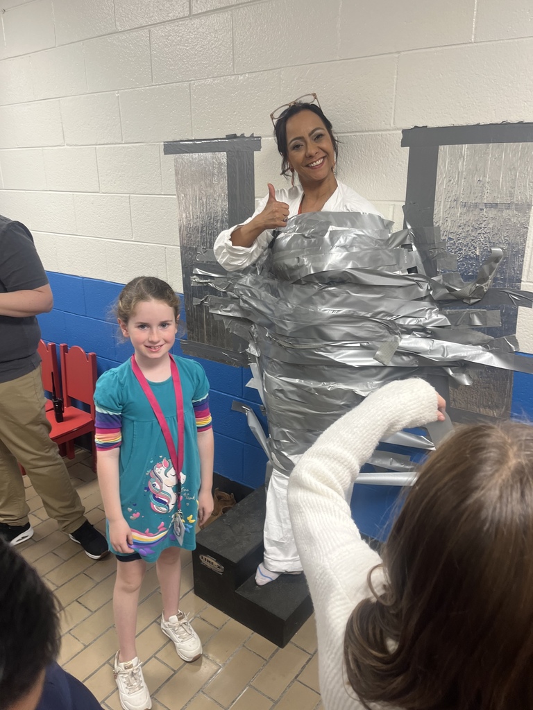 A woman smiles and gives a thumbs-up while taped to the wall, with a young student standing proudly in front of her.
