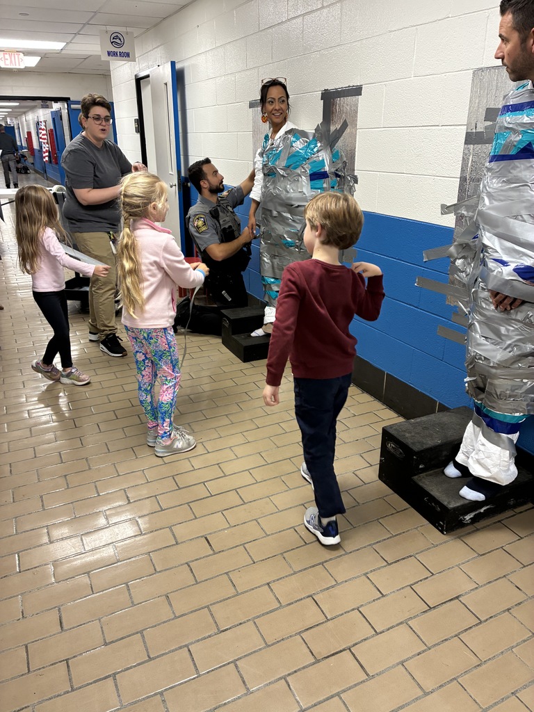 Students and staff continue taping two adults to the wall, with everyone laughing and enjoying the playful school activity.