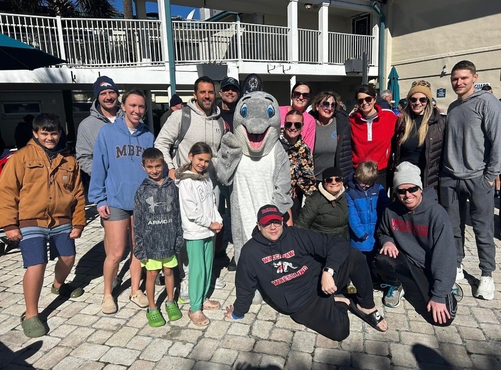A group of adults and children pose together outdoors on a sunny day in front of a beachside building. In the center stands Splash the Dolphin mascot, smiling and waving, surrounded by team members dressed in jackets, hoodies, and winter hats. The group appears cheerful and bundled for cool weather as they gather to support the Polar Plunge.