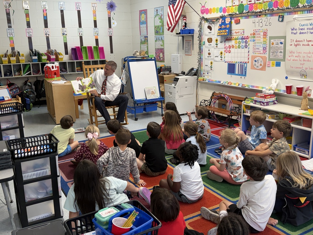 A male guest reader sits in a rocking chair reading to a group of young students gathered on a colorful rug in a classroom.