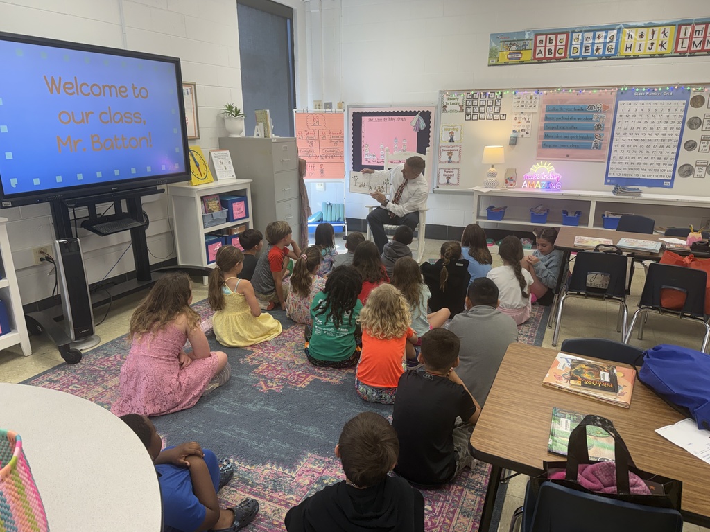 A male guest reader sits at the front of a classroom reading aloud while students sit on the carpet listening; a screen nearby welcomes the visitor.