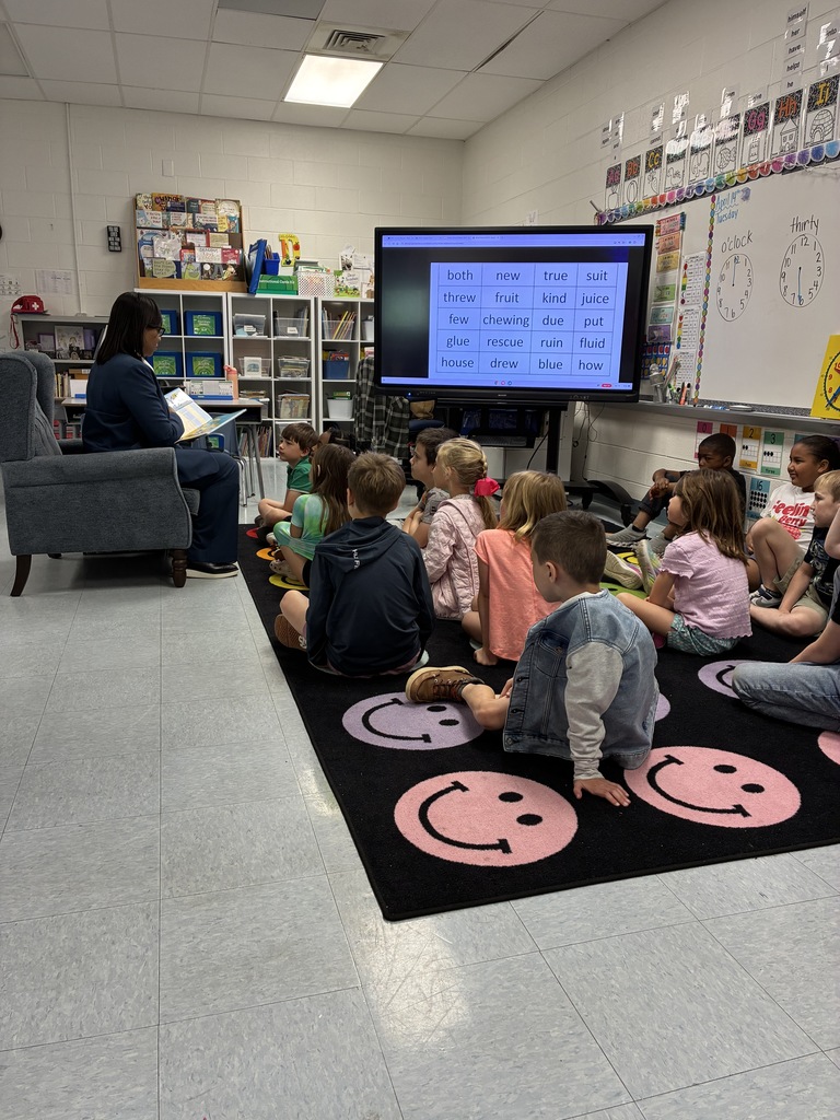 A guest reader sits in a chair reading a book aloud to a group of elementary students बैठे on a colorful classroom rug, with a digital board displaying vocabulary words behind them.