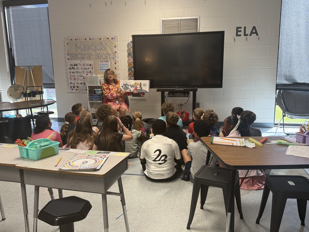 A guest reader holds up a picture book while reading to a group of students बैठे on the floor in an elementary classroom.