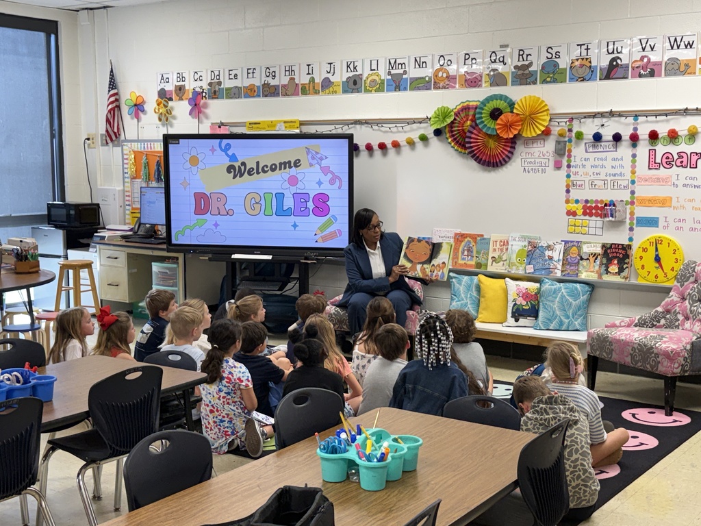 A guest reader sits at the front of the classroom reading a picture book while students sit attentively on the floor; a screen behind her welcomes a special guest.