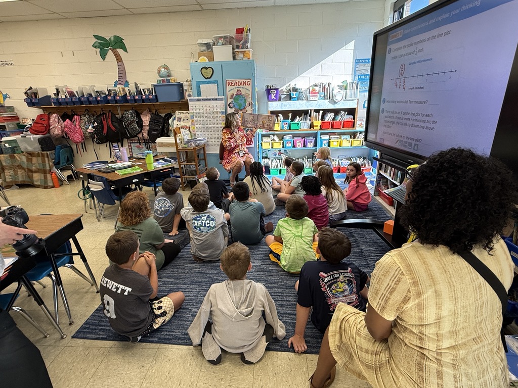 A guest reader shows illustrations from a book to a group of students seated on the floor in a classroom filled with books and learning materials.