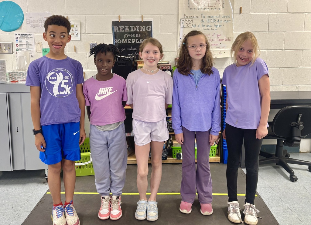 Five students standing together in a classroom wearing shades of purple.