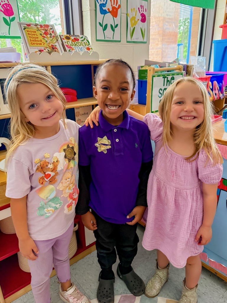 Three young students smiling and wearing purple outfits in a classroom.