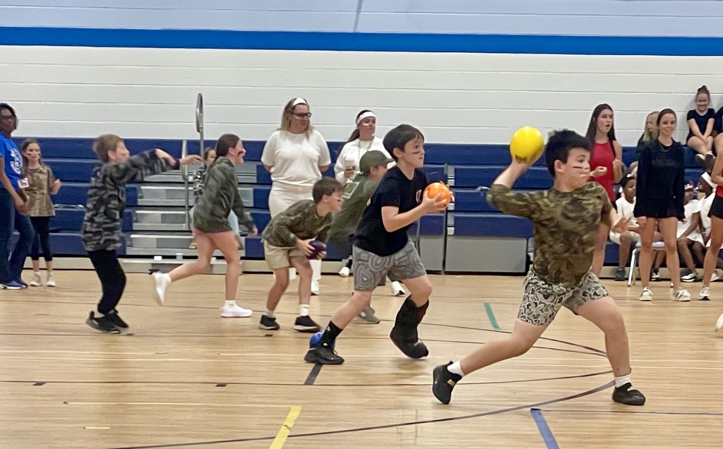 Several elementary students actively play dodgeball in the gym, with one student mid-throw and others preparing to catch or dodge.