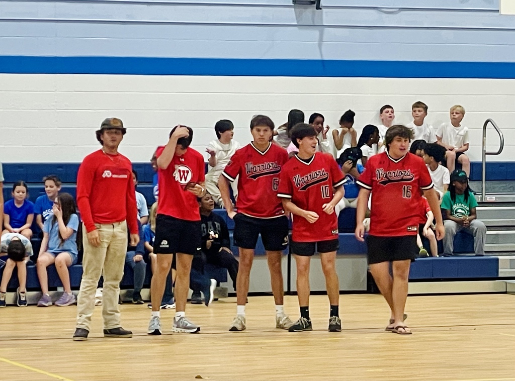 A group of high school baseball players in red uniforms stand together on the gym floor, watching the dodgeball game.