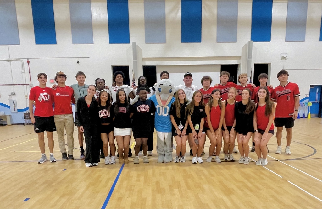 A large group of high school athletes, cheerleaders, and a school mascot pose together in a gymnasium, smiling for a group photo.