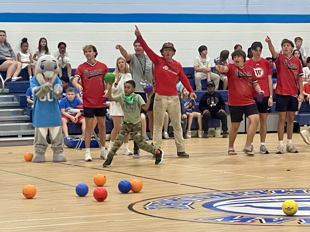 A student throws a dodgeball while high school athletes in red jerseys cheer and point excitedly from the sideline; a mascot stands nearby.