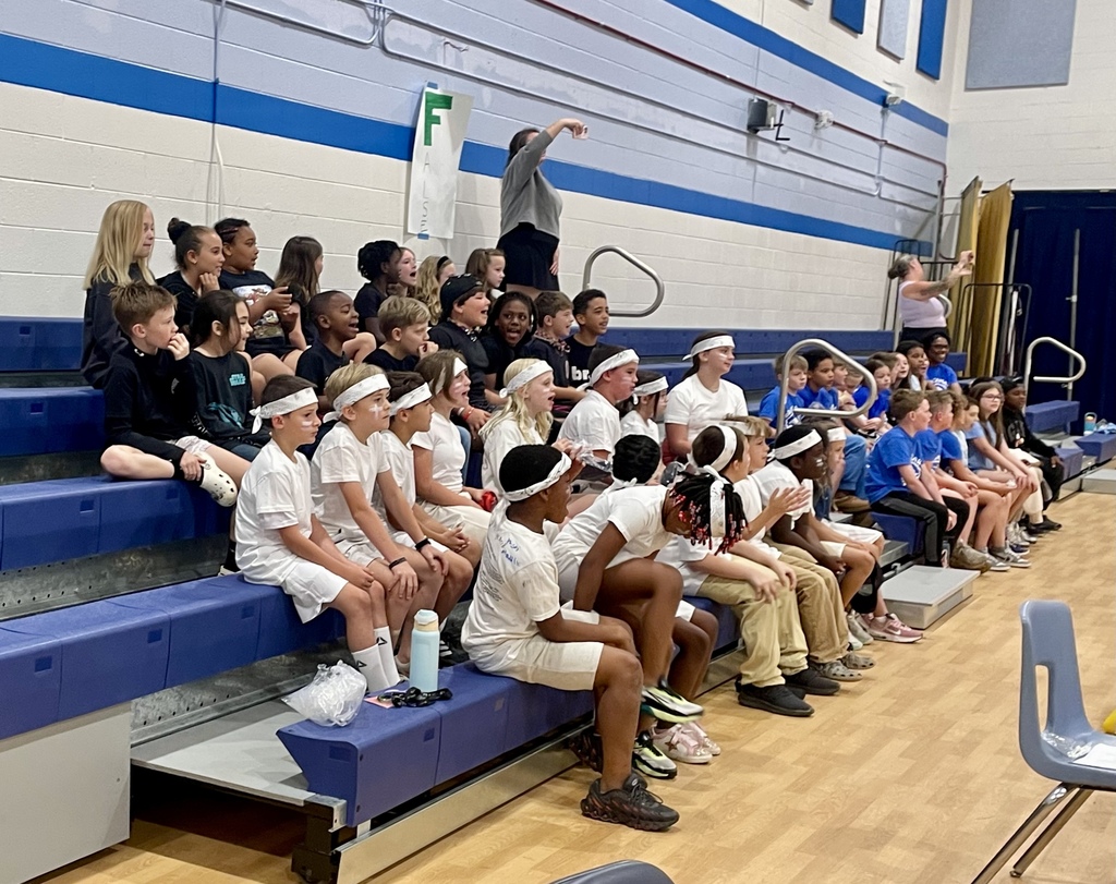 A group of elementary students sit on bleachers, dressed in coordinated outfits, watching and cheering during the pep rally.