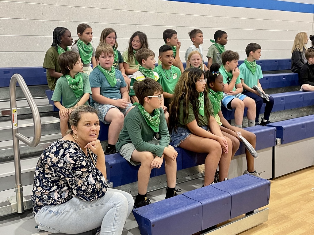Students wearing green bandanas sit together on bleachers, watching the event while a teacher sits nearby.
