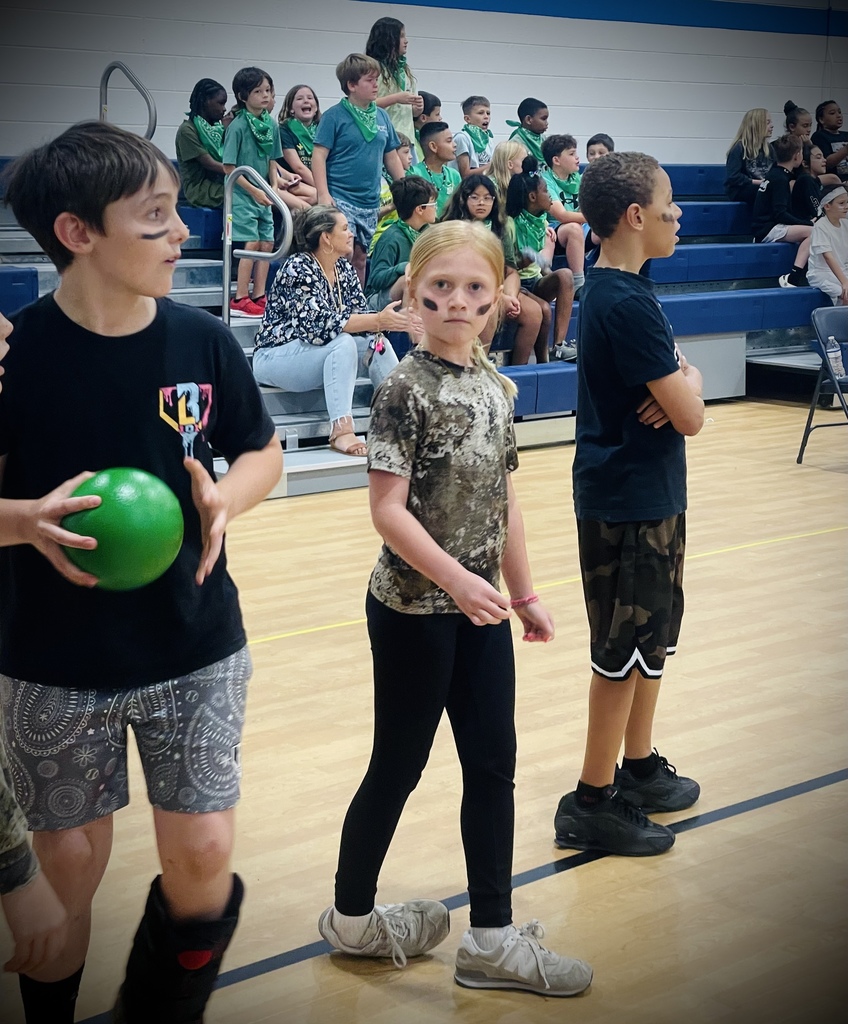 More students in green shirts and bandanas sit and stand on bleachers, reacting and cheering during the pep rally.
