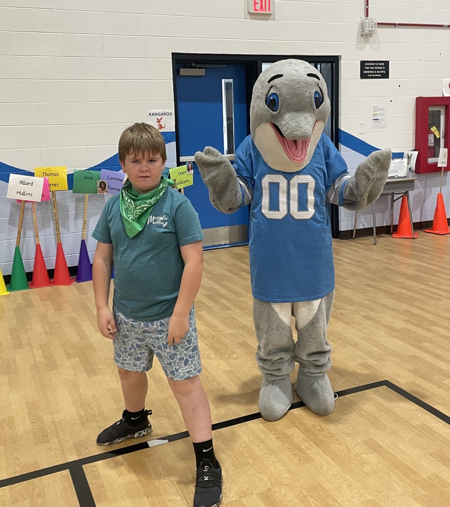 A student stands next to a smiling school mascot in a gym, both facing the camera during the event.