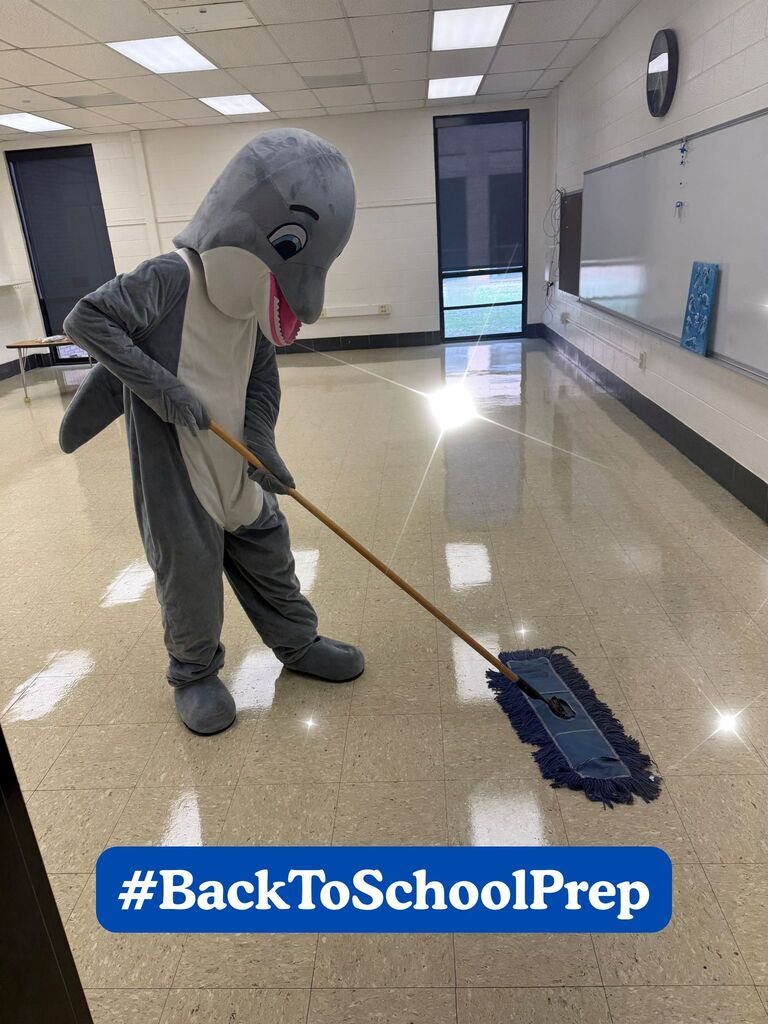 The dolphin mascot carefully dust mops a shiny classroom floor in an empty classroom, with sparkling reflections emphasizing the freshly cleaned space. A #BackToSchoolPrep banner appears at the bottom of the image as part of Waccamaw Elementary's summer preparation series.