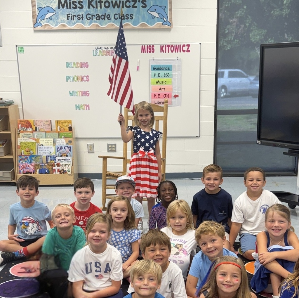 Waccamaw Elementary first grade students dressed in red, white, and blue pose together in their classroom for Patriot Day. One student stands behind the group holding an American flag.