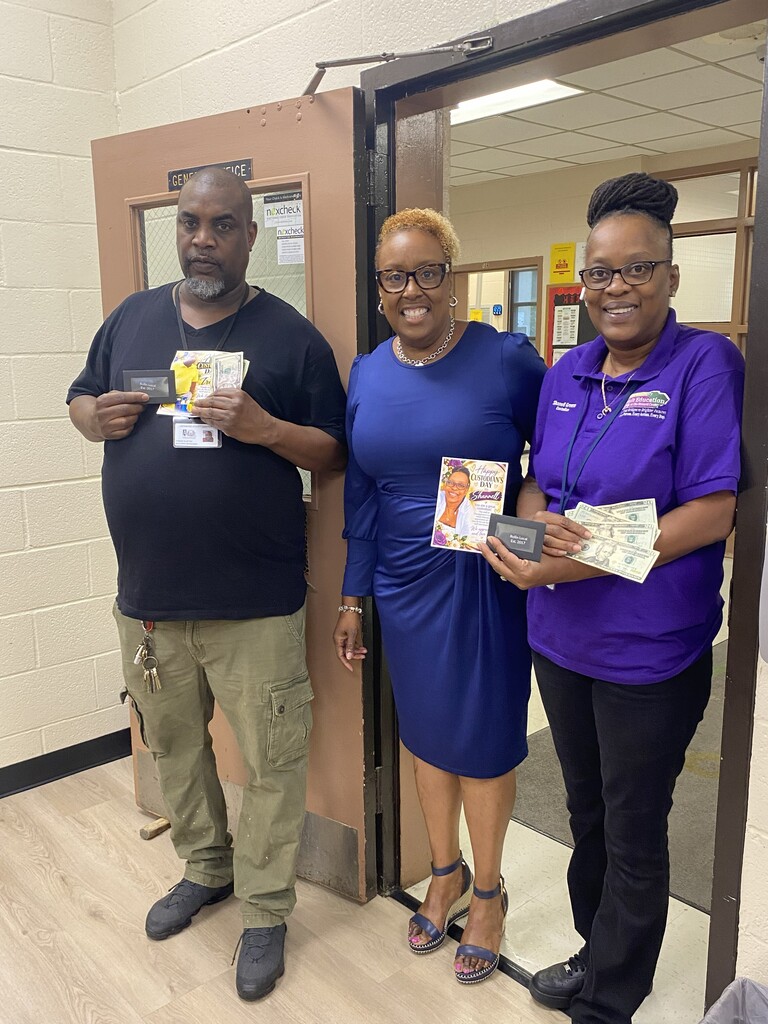 Mr. Travis Alston (left), Dr. Anderson, Director of Georgetown County Adult Education (center), and Ms. Shannell Greene (right) pose together in a school doorway. Dr. Anderson, wearing a dress, holds a "Happy Custodians Day" greeting card while Mr. Alston and Ms. Greene each hold gift cards as tokens of appreciation from the staff for their dedicated service to the school community.