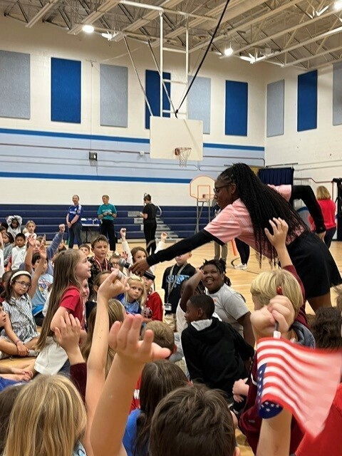 Melissa Jefferson-Wooden interacts with students seated on the gym floor during the school assembly, with many students raising their hands.