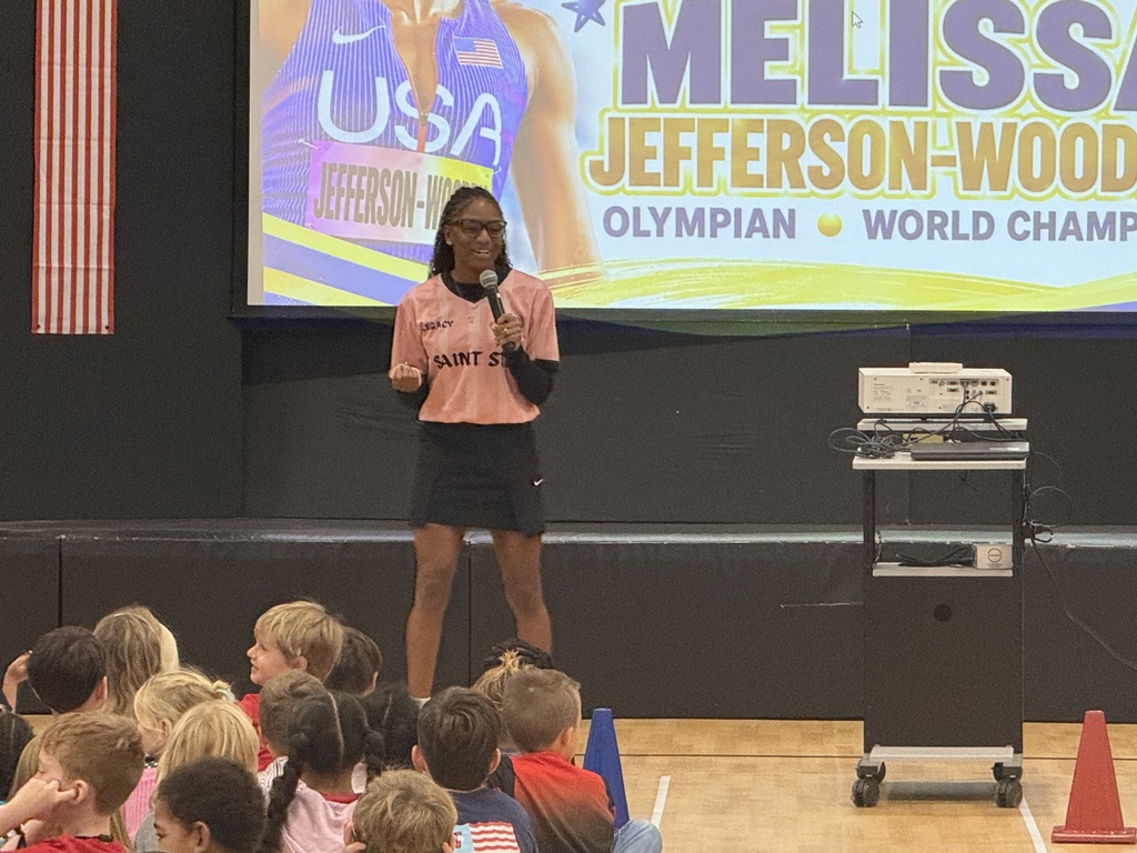 Melissa Jefferson-Wooden speaks into a microphone during the assembly with a welcome graphic displayed behind her.
