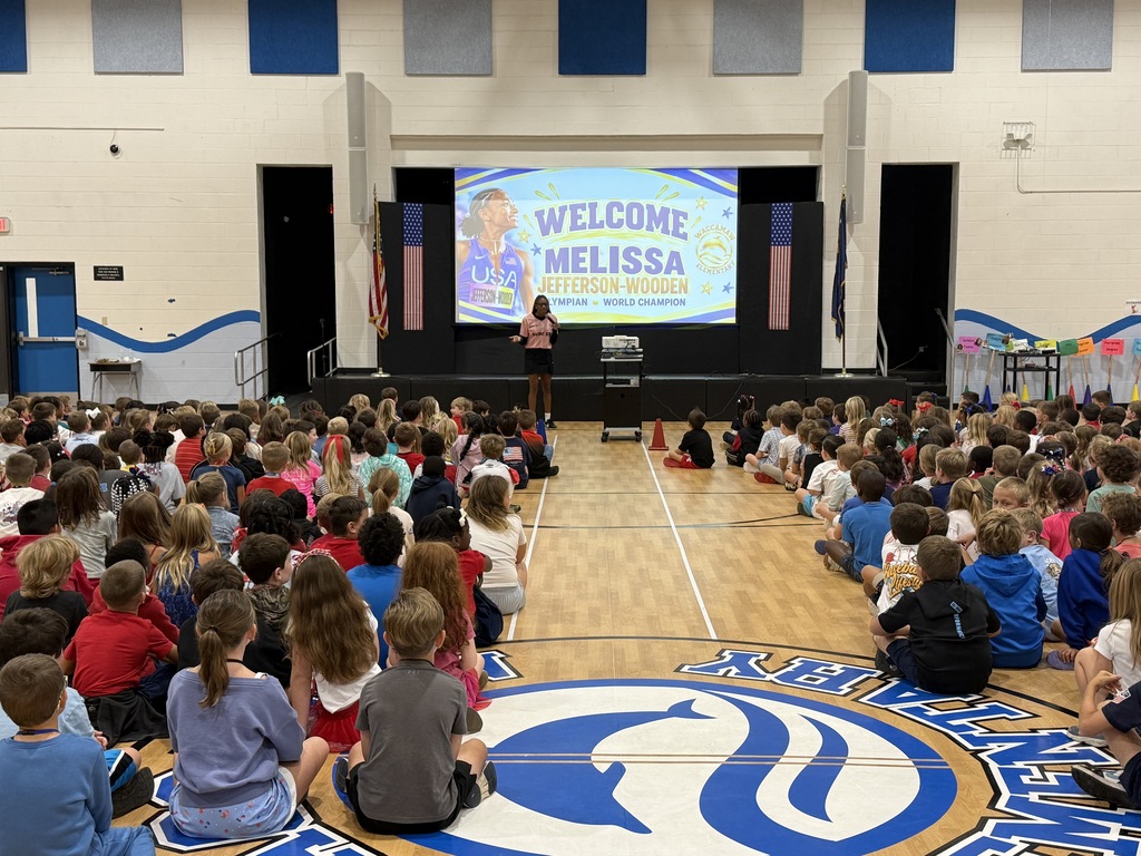 Wide view of the Waccamaw Elementary gym filled with students listening to Melissa Jefferson-Wooden speak beneath a projected welcome graphic.