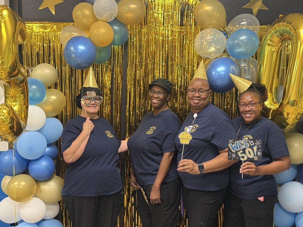Four staff members in navy shirts pose in front of the gold and blue anniversary photo backdrop, holding celebration props.