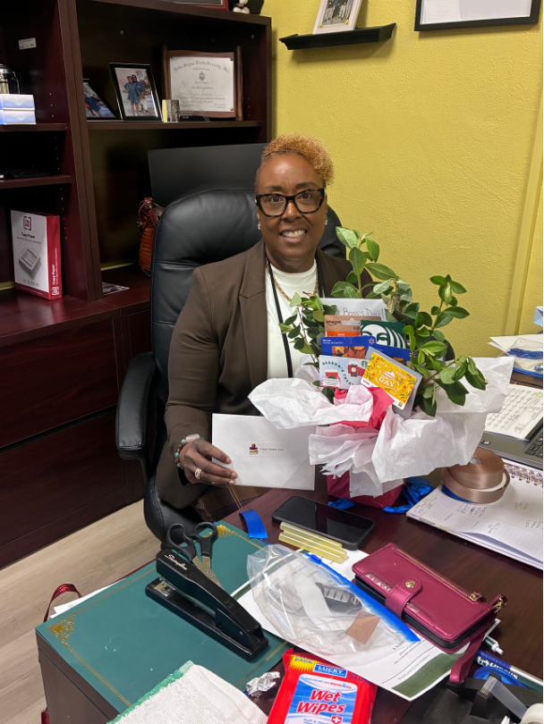 A moment with Dr. Anderson at her desk—grateful for her commitment to education, student advocacy, and our GCAE community. Dr. Paula Anderson, who has short light-colored hair and glasses, sits smiling at her office desk holding a wrapped gift basket with green plants, tissue paper, and cards. Shelves and framed documents are visible behind her.