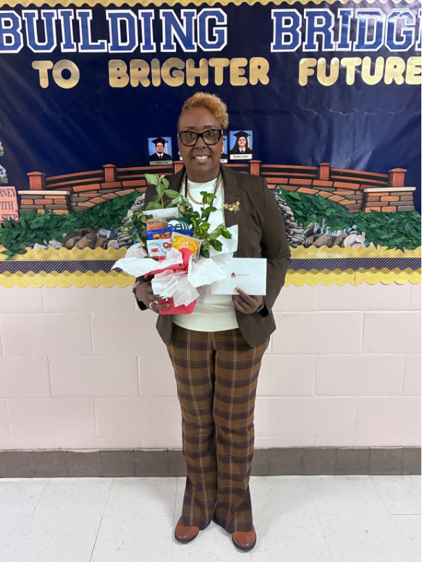 Dr. Paula Anderson stands holding a bouquet of flowers and a white card in front of the lobby display, which shows a brick bridge over greenery, the words “Building Bridges to Brighter Futures,” and two small graduation portraits.