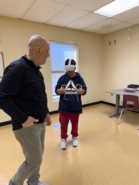 A student wearing a white virtual reality headset with controllers. The students is participating in a virtual career experiences, for example knee replacement surgery . The student is wearing red pants and blue and white hoodie. The adult that is assisting the student is wearing khaki pants and navy blue sweater on the left.