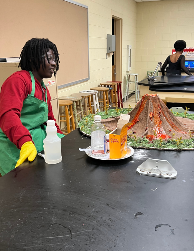 Student wearing a red top and green apron, stands over a volcano. that he made. Therevare liquid items on the table long with the volcano