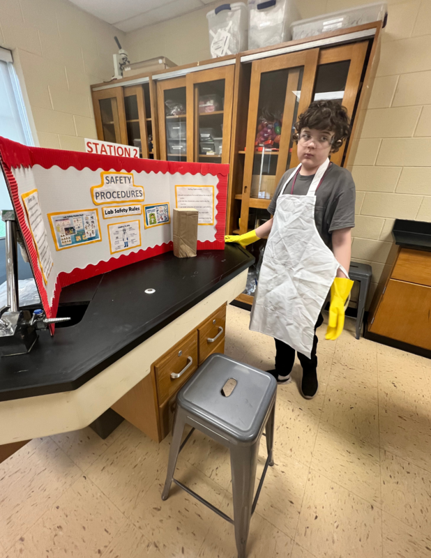 A student standing by his showcase exhibit. He is wearing a gray shirt with a white apron. His project  is on Safety Procedures.