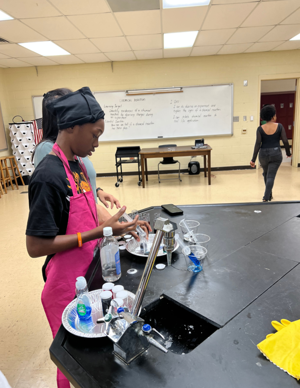We have another student who is wearing a black shirt and red apron and a black head wrap on her head.  She has several liquids in bottles around her, on a table,  in the science lab. She is doing some type of experiment.