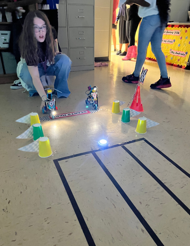 A female student wearing blue jeans and a dark shirt. She is in the STEM classroom and she is preparing to race two Lego robots on bikes in the Smart Bike Showcase. There is a racetrack made of lines  on the floor. 