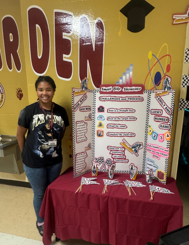 A female student wearing blue jeans and a black t-shirt. She is standing by the STEM exhibit called  Smart Bike Showcase. The Challenges and Process are to guide  the Lego robots in a  straight line, stop at the target area, climb the ramp, and finish the race.