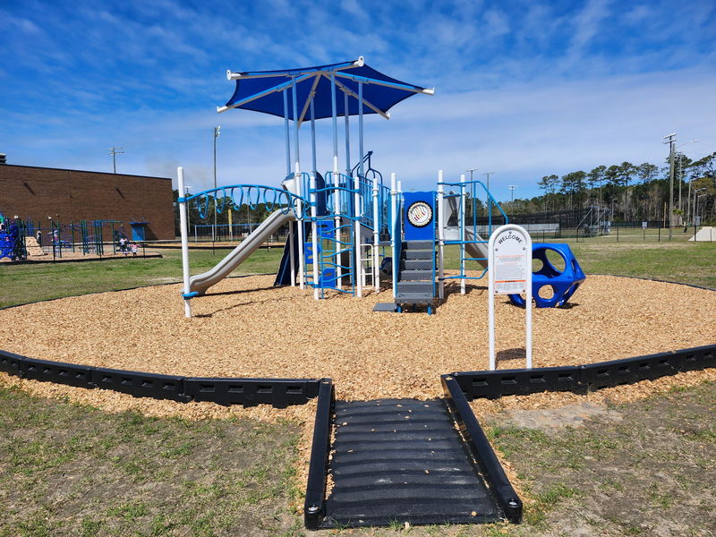 “New playground structure with slides, climbing features, and a blue shade canopy, set on a wood chip surface with a ramp entrance, located on a school playground with fields and trees in the background.”