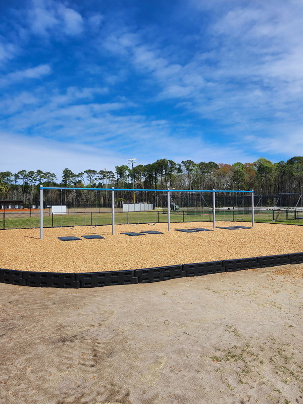 “New playground swing set with multiple swings over a wood chip surface, enclosed by a black border, with open fields and trees in the background under a blue sky.”