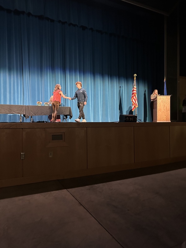 Spring 2026 Academic Awards Ceremony. A student crosses the stage to receive an award from school administrators after their name is announced by Student Services staff.
