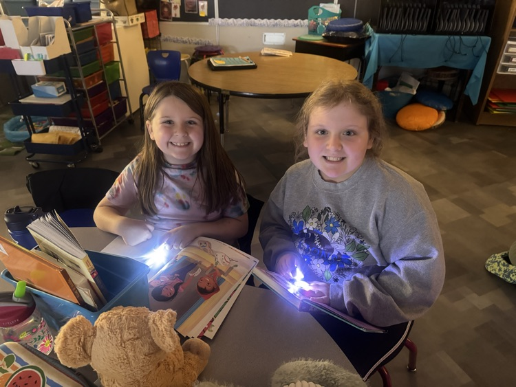 two students reading at a desk using flashlights 