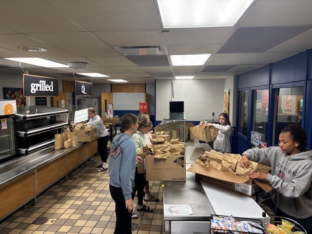 Students are in the cafeteria organizing breakfasts into boxes for distribution 