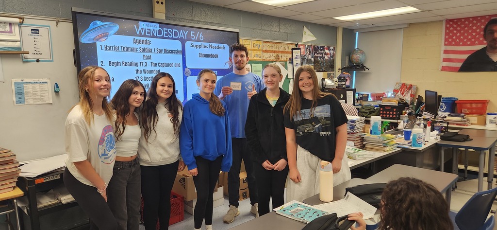 Sixth students stand with a teacher in front  of a digital board smiling for the camera