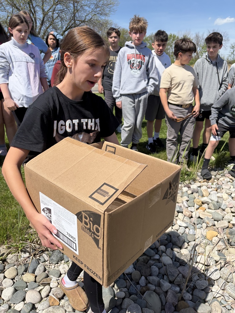 A student is carrying a box that is partially closed containing baby ducks to the pond while students look on from behind her.