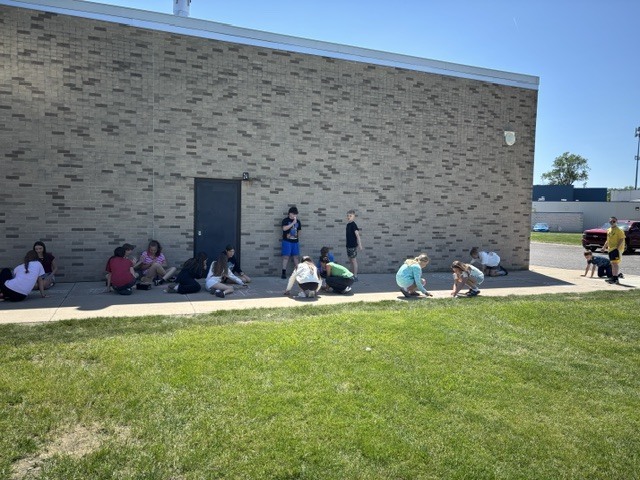 Students are scattered around outside against the side of the builing drawing on the sidewalk with chalk