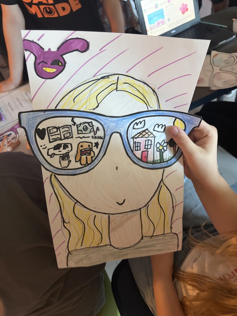 A picture of a girl with glasses on held by a hand with a computer in the background
