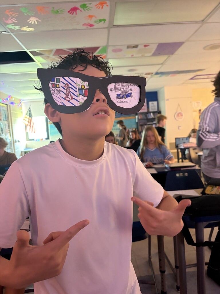 A student with a pink shirt wearing paper glasses and pointing at his face in a classroom with students in the background